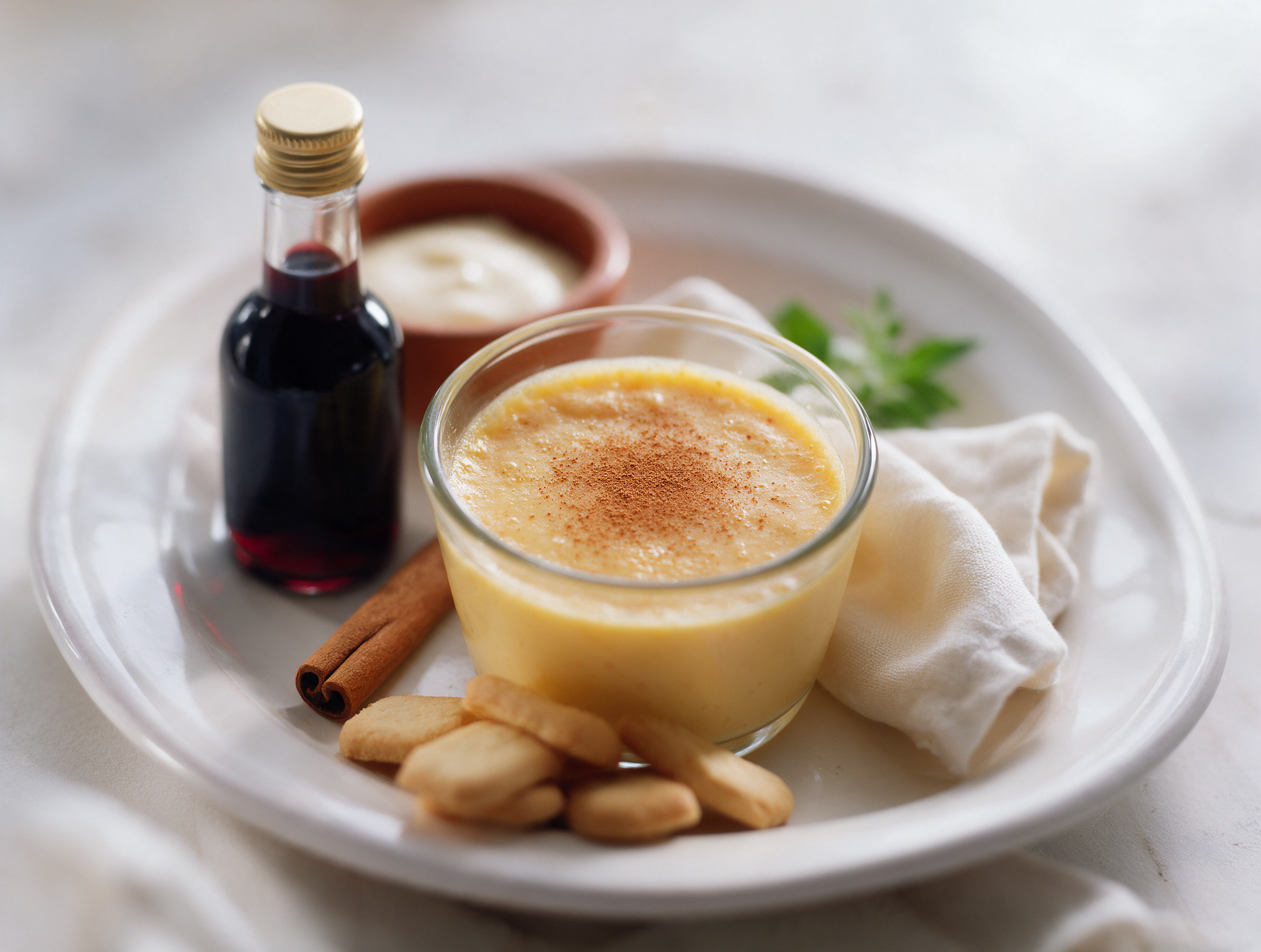 Pale golden Rüsümada Valtellinese in a small glass cup dusted with cinnamon, wine bottle resting on a rustic wooden table.