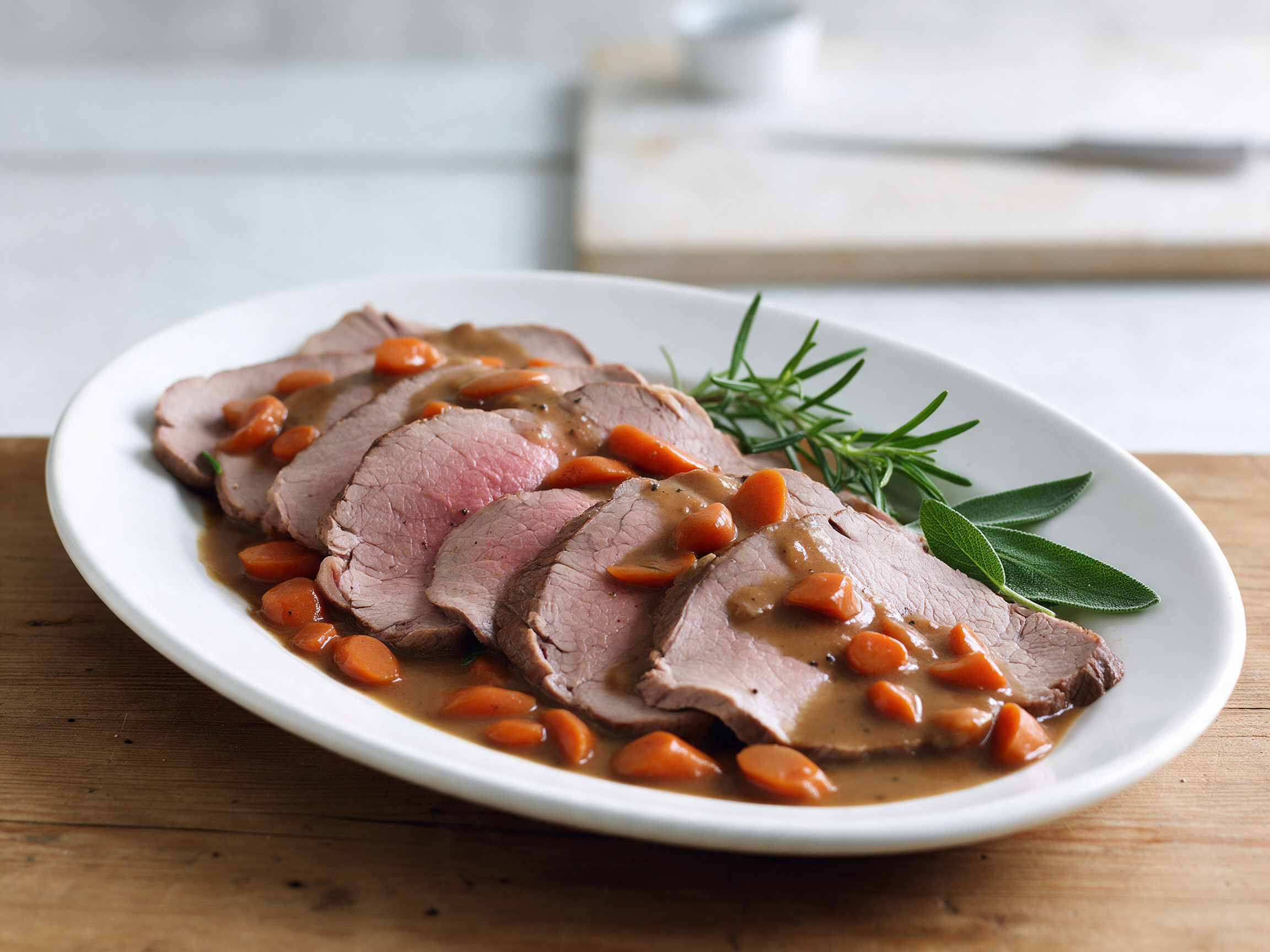 Sliced vitello arrosto fanned on a white platter with rosemary pan sauce, carrots and sage on a wooden table